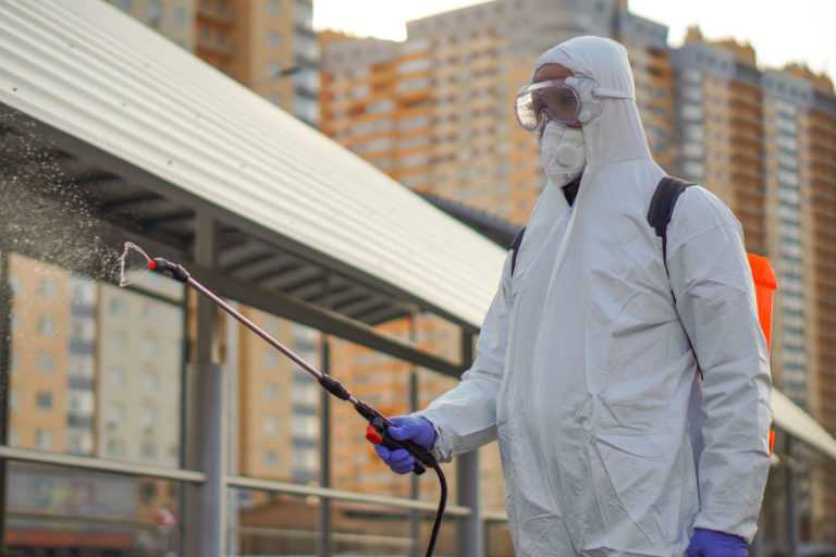 A man in protective equipment disinfects with a sprayer in the city. Surface treatment due to coronavirus covid-19 disease. A man in a white suit disinfects the street with a spray gun. Virus pandemic