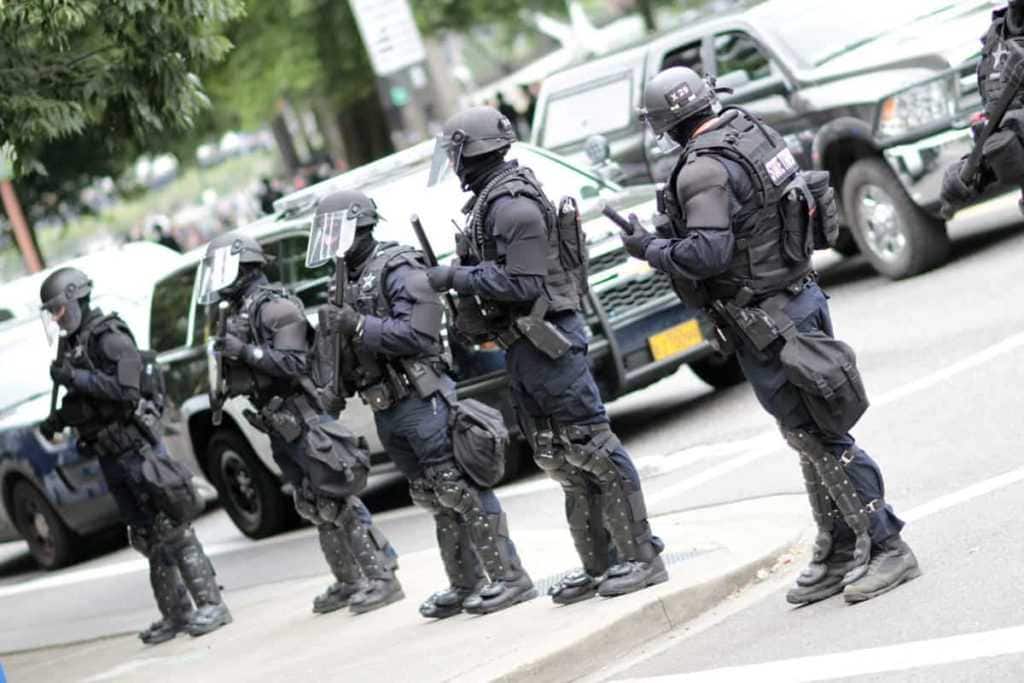 Police officers in riot gear overseeing a crowd of protestors.